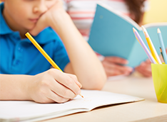 Student writing in a notebook and a student reading a notebook as they sit at a desk with a cup filled with pencils in it